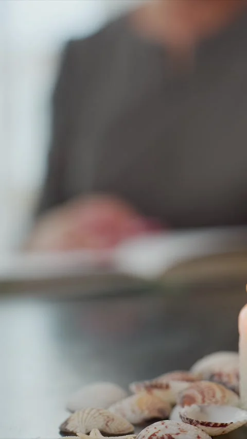 Close up of lit candle and seashells on smooth surface with person reading book Stock Footage 345728614