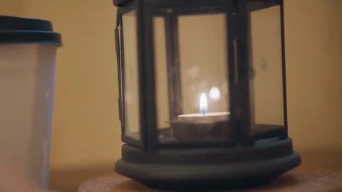Close up of lit candle in glass lantern beside white cup on wooden table with Stock Footage 346729449