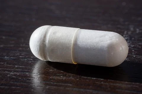 Close-up macro shot of a white capsule pill resting on a dark wooden surface. Stock Photos