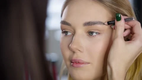 Close-up of a make-up artist drawing eyebrows with an eyebrow pencil on a young Stock Footage 235659579