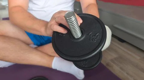 Close-up of a man assembling a black cast iron dumbbell while sitting on a pu 库存照片