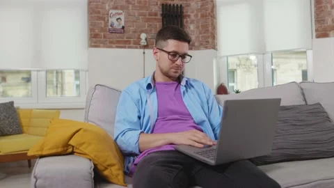 Close up man freelancer sitting on sofa use laptop shrugs at home online working Vidéo 133791098