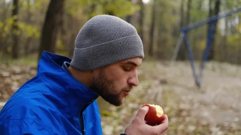 Close-up of man in grey beanie and blue jacket eating red apple in autumn park Stock Footage 315584904