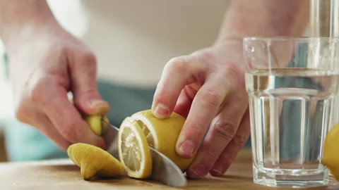 Close up of man hands slicing fresh lemon on wooden board near glass of water, h Stock Footage 332680439
