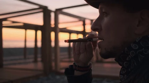 Close-up, a man in a hat and leather jacket Smoking a cigar at sunset . 50 fps Stock Footage 121806257