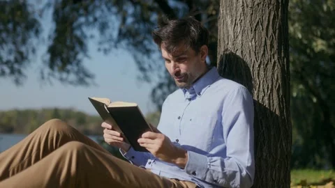 Close up of man reading book and talking while leaning on park tree Stock Footage 332298936