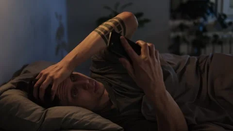 Close-up of man scrolling on phone in bed at night, shocked by what he sees. Stock Footage 296057808