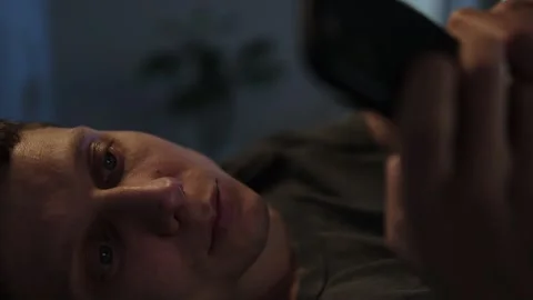 Close-up of a man scrolling through his phone in bed at night, struggling with s Stock Footage 296423025