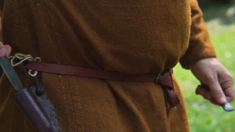 A close-up of a man in simple medieval clothing sharpening a knife and putting Vídeo Stock 345033680