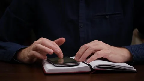 Close-up. man sits at desk with diary and takes notes in smartphone. front view. Stock Footage 303137451