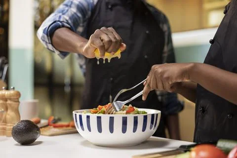 Close up of man squeezing lemon juice over produce and woman mixing veggies Fotos Stock