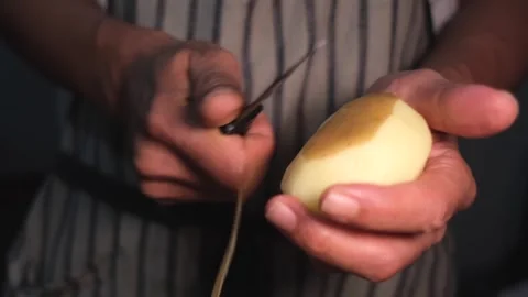 Close up a man in a striped apron peeling raw fresh a potato with a knife. Stock Footage 247546754