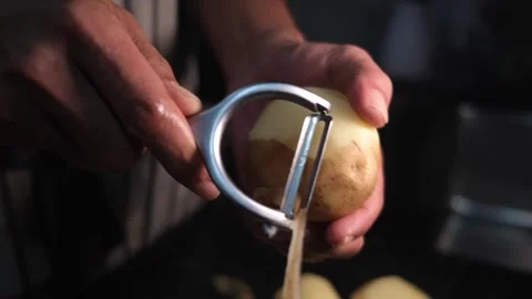 Close up a man in a striped apron peeling raw fresh a potato with a peeler. Stock Footage 247546973