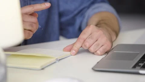 Close-up of a man at a table in front of a laptop making notes in a notebook. Stock Footage 236588650