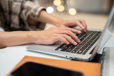 Close up of a man's both hands typing on laptop with phone atop book sittin.. Stock Photos