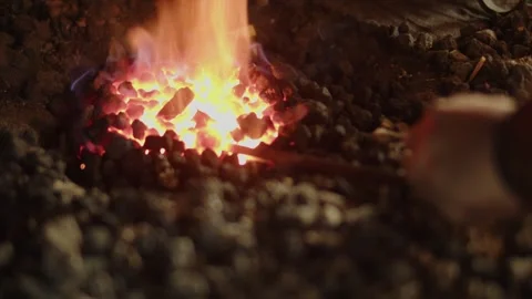 Close-up of a man’s hand turning a metal rod in flames among charred rocks and Video stock 336428535