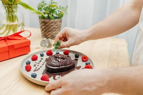 Close-up of a man's hands decorating a chocolate heart cake with a sprig of Stock Photos