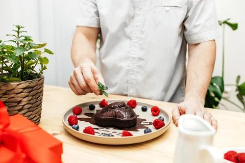 Close-up of a man's hands decorating a chocolate heart cake with a sprig of Stock Photos