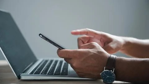 Close-up of a man's hands using a smartphone with a laptop in front of him... 库存照片