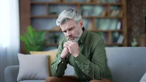 Close-up of mature bearded man with gray hair sitting alone on sofa at home look Stock Footage 328707010