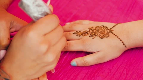A close-up of a mehendi master draws henna on person's hand on a pink background Stock Footage 240719556