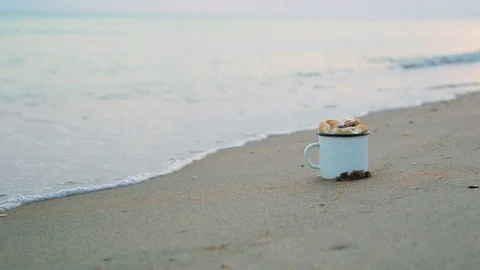 Close-up mug filled with shells stands on sandy shore against blurred sea wave 動画素材 93110662