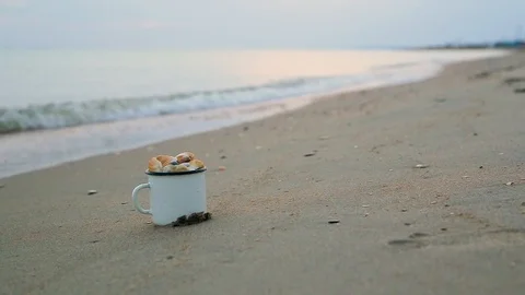 Close-up mug filled with shells stands on sandy shore against blurred sea wave Stock Footage 93110707