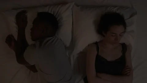 Close-up of multiracial couple lying in bed at night. Caucasian girl sights and Stock Footage 136576732