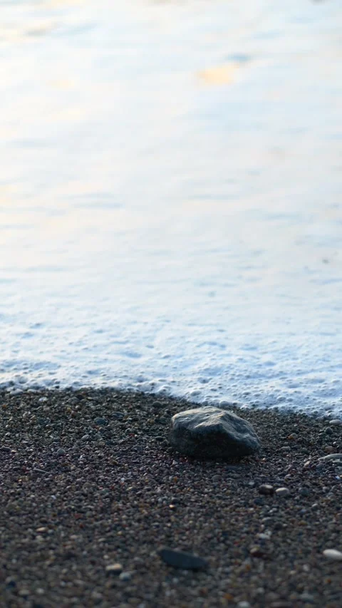 Close-up of ocean waves rolling over a small stone on the shoreline, with white Stock Footage 332896478