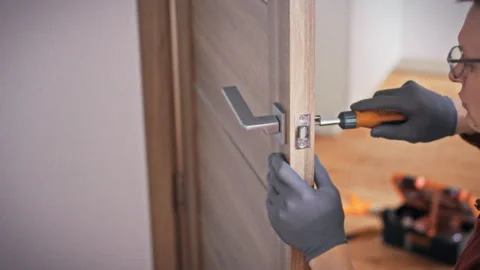 A close-up of an orange screwdriver in gloved hands working on a door lock m 動画素材 344877147