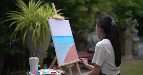 Close up of painting tools and canvas showing pink and blue seascape with woman Stock Footage 333602072