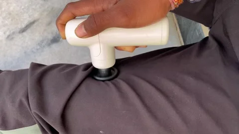 Close up of a person using a white portable electric massage gun on their leg Видео 328352286