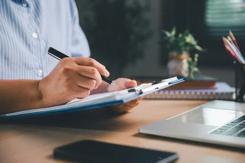 Close-up of a person writing on documents attached to a clipboard beside a .. Stock-Fotos
