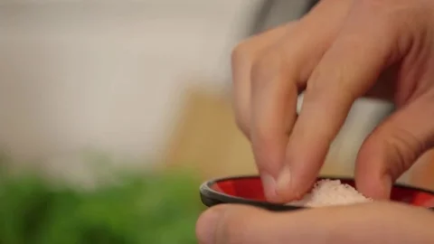 Close up pinch of salt in the kitchen, defocussed herbs in background Video stock 74605560