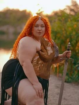 Close-up Plus Size Redhead Warrior Woman Kneeling Holding Sword by Lake Stock Photos
