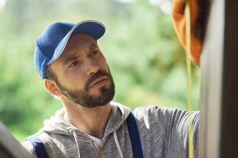 Close up portrait of focused young builder wearing blue cap and overalls using Foto stock