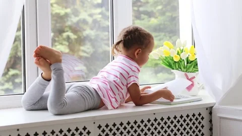 Close up portrait of little cute girl reading book on windowsill at home. Stock Footage