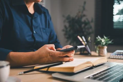 Close-up of a professional using a smartphone above an open book and laptop.. Stock Photos