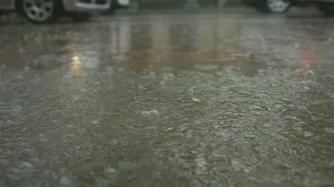 Close-up of raindrops falling into a puddle on the pavement, cars driving in the Stock Footage 204948677