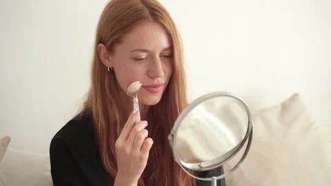 Close-up of redhead woman using jade roller on face looking in the mirror. Stock Footage 171931540