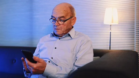 Close up of retired man sitting at home with his mobile phone at night Stock Footage 99014615