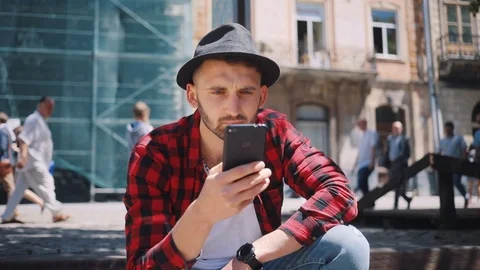 Close up serious handsome young man in the hat uses phone sitting in city Stock Footage 92071753