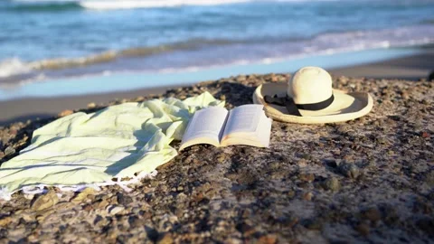 A close up on a shawl, straw hat, open book, sandy beach with blue waves. Stockbeeldmateriaal 134655269