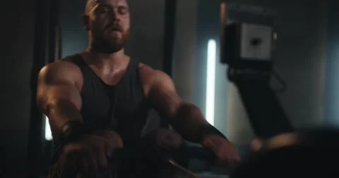 Close-up shot of a bearded man in a tank top exercising on a rowing machine in a Stock Footage 313358196