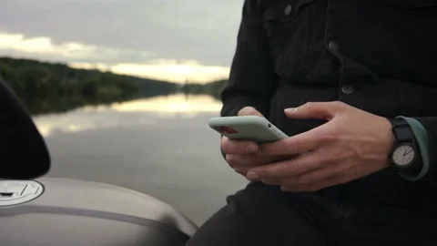 Close-up shot of man messaging on the phone sitting on motorbike by the lake Video stock 162855463