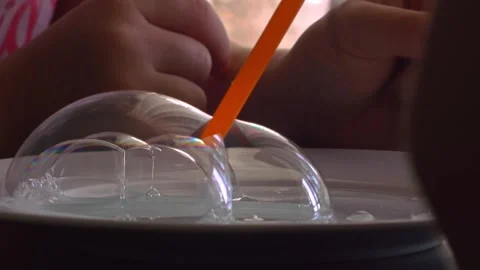 Close-up shot of a orange straw blowing soap bubbles on a tabletop, with a Video stock 328912868