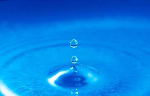 Close-up of a single water drop falling into rippling blue liquid surface Stock Photos