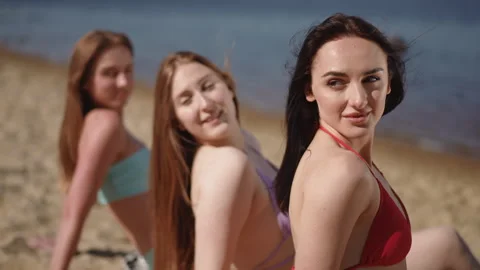Close-up. Sitting on a sandy beach, three bikini girls sunbathing look over Stock Footage 267056769