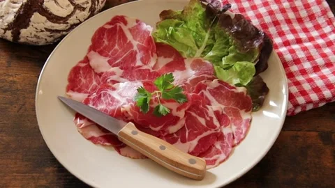 Close up of slice of coppa in a plate with salad and a ball of bread on a table Stock Footage 177914134