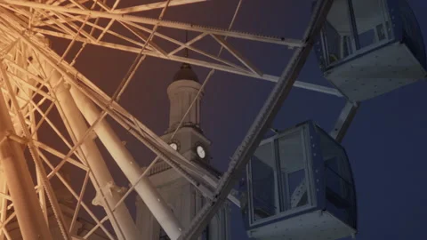 Close up of slowly moving ferris wheel against a clock tower, evening sky Stock Footage 259386589